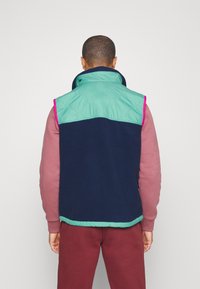 Marineblaue Fleece-Weste mit einem türkisfarbenen gesteppften Oberteil, pinkem Besatz und hohem Kragen, getragen über einem hellrosa Langarmshirt.