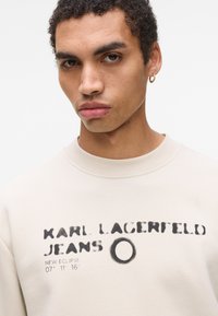 Bežinė megzta palaidinė su apykakle, su juodu spausdintu tekstu "KARL LAGERFELD JEANS" ir apvaliu dizainu. Minkšta tekstūra, laisvas siluetas.