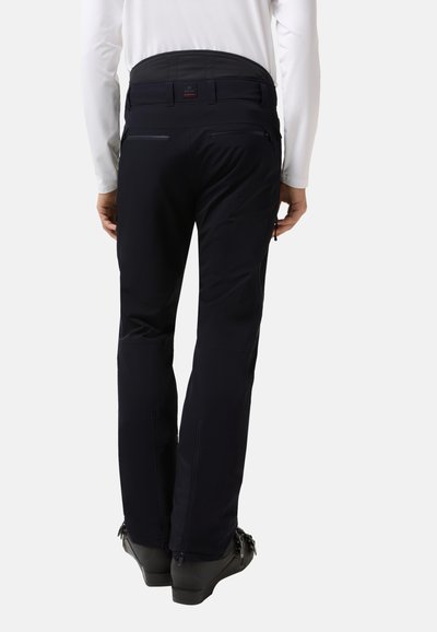 Osoba ve černých lyžařských kalhotách a černých lyžařských botách, zobrazená zezadu na jednobarevném bílém pozadí.