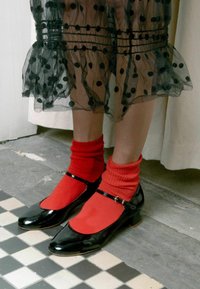 Zapatos Mary Jane de charol negro con punta redonda, acentuados por una correa delgada y un hebilla, combinados con calcetas rojas brillantes con textura de canalé.