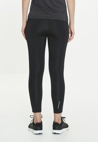 Des leggings noirs en matériau lisse et extensible avec un détail à la cheville composé de trois petits points blancs. Portés avec des chaussures de sport noires.