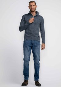 Suéter de punto azul marino con cuello alto y ribete acanalado, combinado con jeans azul claro y botas negras con cordones, de pie contra un fondo liso.