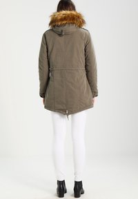 Femme portant un parka vert olive avec une capuche bordée de fourrure, un pantalon blanc et des bottines noires, debout de dos devant un fond uni.