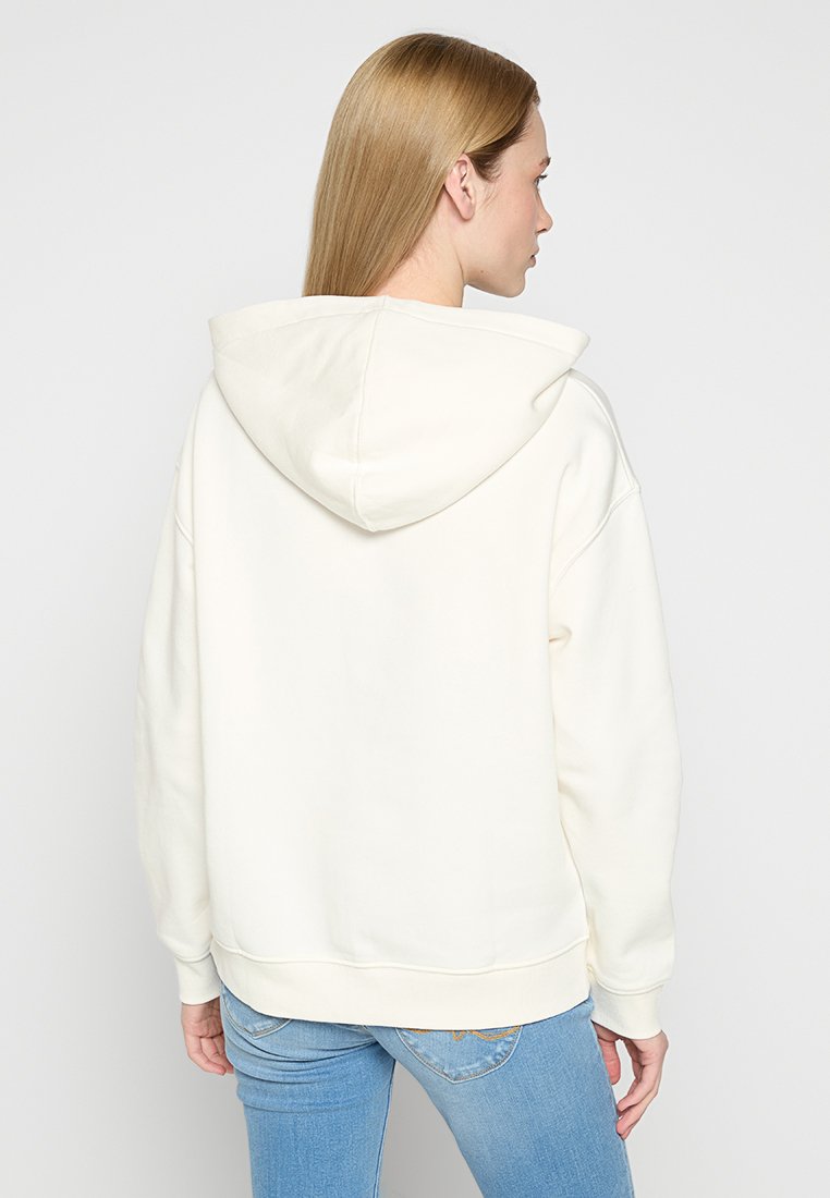 Sudadera de color crema con una textura suave, que presenta una capucha con cordones, puños acanalados y un corte relajado, vista desde la parte posterior.