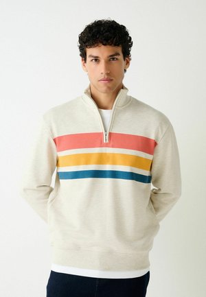 Junger Mann mit lockigem dunklem Haar, der einen beigen Pullover mit roten, gelben und blauen horizontalen Streifen und einem Viertel-Reißverschluss trägt und vor einem weißen Hintergrund steht.