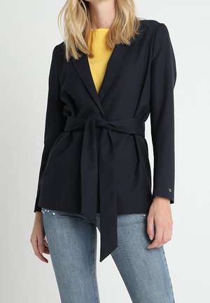 Femme portant un blazer noir ceinturé par-dessus un haut jaune et un jean bleu clair avec des détails en perles près des poches.
