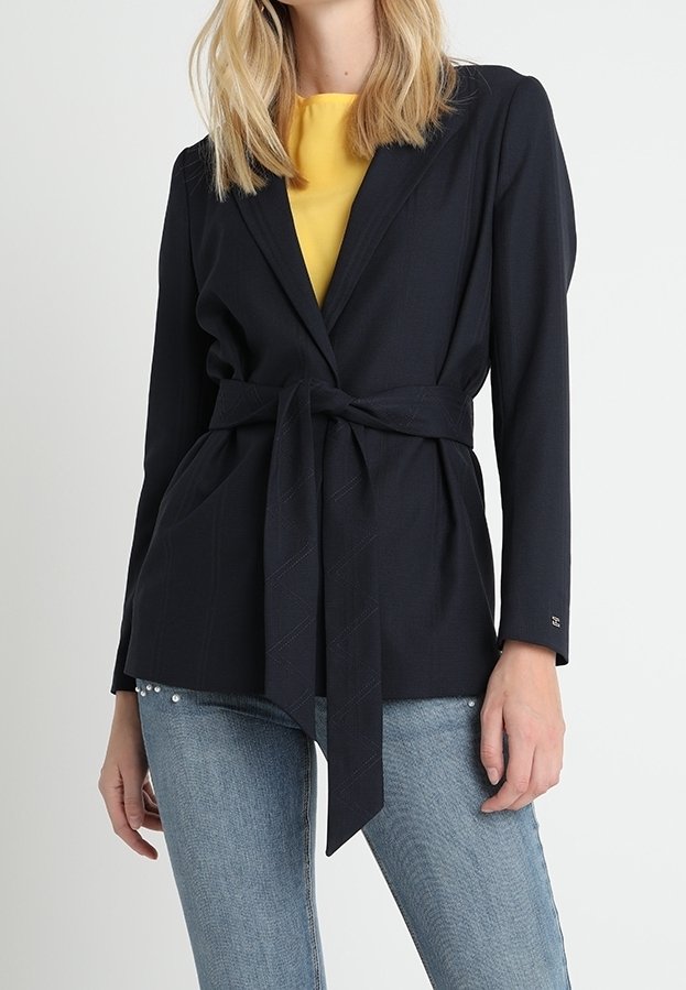 Femme portant un blazer noir ceinturé par-dessus un haut jaune et un jean bleu clair avec des détails en perles près des poches.