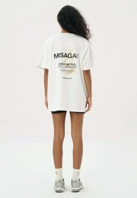 Wit oversized t-shirt met zwarte en gouden tekst op de achterkant. Gepaard met zwarte shorts en zilveren sportschoenen. Eenvoudige achtergrond.