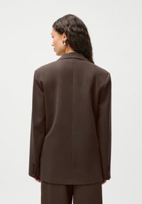 Brun skreddersydd blazer med en skarp krage, glatt tekstur, enkelt knappefeste og lange ermer. Enkel design med minimalistiske detaljer.