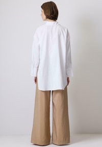 Camicia bianca oversize con maniche lunghe, tessuto strutturato e orlo irregolare, abbinata a pantaloni beige a vestibilità ampia.