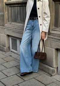 Leveled blue-faded wide-leg jeans dengan pinggang tinggi, dipasangkan dengan mantel berwarna beige muda dan tas tangan suede coklat tua. Kain berpola dan garis yang bersih.