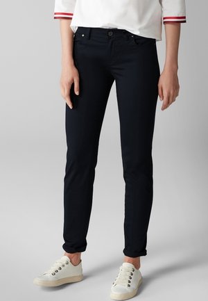 Kobieta nosząca czarne, dopasowane spodnie, białe sneakersy oraz białą koszulę z długim rękawem z czerwonymi paskami na mankietach, stojąca na szarym tle.