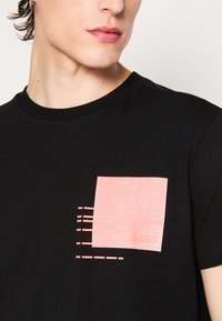 T-shirt noir avec un graphique rose texturé sur la poitrine présentant des motifs rectangulaires et linéaires. Manches courtes et design à col rond.