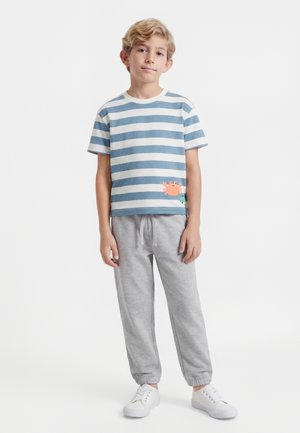 Niño con camiseta de rayas azules y blancas con diseño de cangrejo, pantalones deportivos grises y zapatillas blancas, de pie contra un fondo blanco.