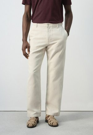 Hombre con pantalones rectos color crema, camisa burdeos y sandalias de cuero trenzado beige, de pie contra un fondo liso y claro.