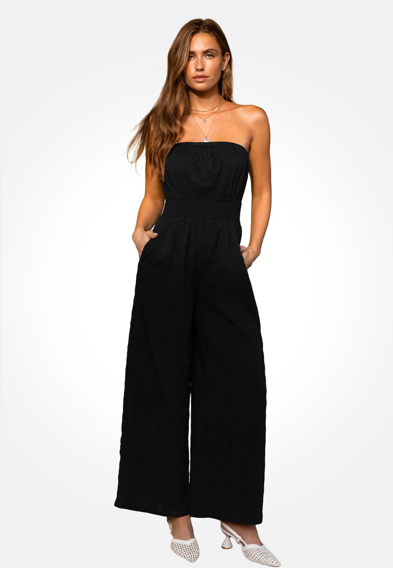 Combinaison noire sans bretelles avec jambes larges et taille élastique, en tissu texturé. Le modèle porte des chaussures blanches à talons et des colliers superposés.