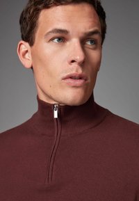 Bordeauxfarbener Zip-Pullover mit hohem geripptem Kragen, ausgestattet mit einem silbernen Reißverschluss und glatter Textur, die ein figurbetontes Design zeigt.