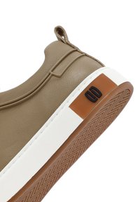 Zapatilla sintética beige con un acabado texturizado, punta redonda y una mediasuela blanca. Presenta un acento marrón en el talón y una suela con diseño.