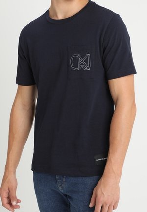 Hombre con una camiseta azul marino oscura lisa de manga corta con un pequeño bolsillo y un logo en el pecho izquierdo y vaqueros azul claro.
