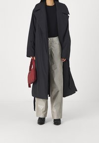 Gabardina negra oversize con solapas amplias, llevada sobre una blusa negra y pantalones plateados brillantes con estampado de cocodrilo. Sostiene un bolso pequeño rojo. Botas negras.