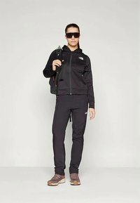 Γυναίκα φορώντας μαύρο αθλητικό σετ με γυαλιά ηλίου και σακίδιο, στέκεται μπροστά σε ουδέτερη φωτεινή φόντο, κοιτώντας μπροστά.