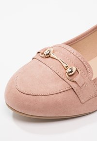 Mocassin en daim rose clair avec un accent de ferrure dorée et des détails de couture. Bout arrondi avec une semelle plate et une texture lisse.