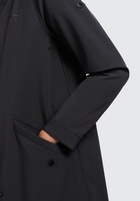 Cappotto impermeabile nero con una texture liscia, dotato di chiusura con bottoni, due tasche laterali e una leggera lucentezza.