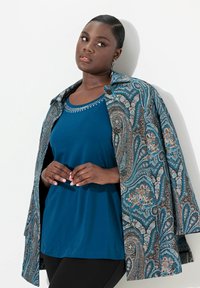 Giacca blu con motivo paisley, chiusa con bottoni, sopra una maglia blu unita. I pantaloni neri e gli orecchini mettono in risalto l'outfit. Tessuto con una texture liscia.