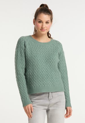 Jonge vrouw met paardenstaart, een gestructureerde groene gebreide trui en lichtgrijze spijkerbroek, staand tegen een witte achtergrond.