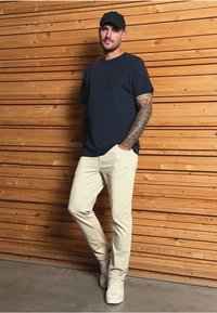 Camiseta de manga corta azul marino, jeans ajustados color crema, zapatillas blancas, brazo tatuado, de pie contra una pared de madera.