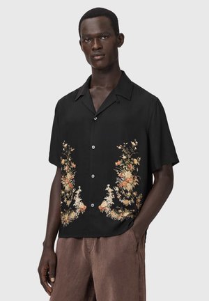 Férfi fekete, rövid ujjú, gombos ingben, krém és narancssárga virágmintákkal, barna nadrággal, kezét részben zsebre dugva.