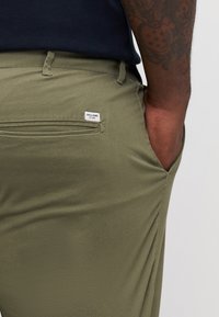 Olive green pants featuring a back pocket with a small logo tag, made from a smooth fabric with a fitted design and subtle texture.