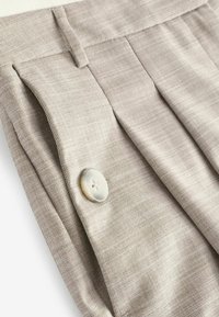 Beige textuurbroek met voorplooien en een grote, ronde witte knop. De stof heeft een geweven patroon en een gladde afwerking.