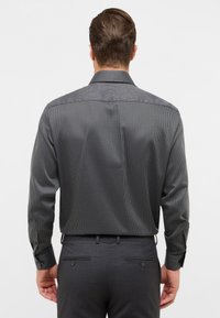 Man in een donker gepatterneerd overhemd met lange mouwen, in zijn donkere op maat gemaakte broek gestoken, gezien van de achterkant tegen een effen achtergrond.
