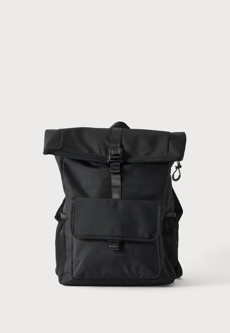 Sac à dos noir à enroulement supérieur avec poche avant à rabat, fermeture à boucle, poches latérales en filet et un crochet sur le côté droit sur un fond uni.