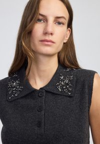 Kobieta z długimi brązowymi włosami, ubrana w bezrękawnik w ciemnoszarym kolorze z ozdobnym kołnierzykiem i czarnymi guzikami, na neutralnym tle.