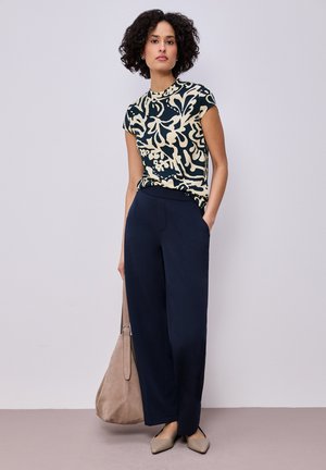 Kobieta o kręconych ciemnych włosach, ubrana w wzorzysty top z krótkim rękawem, granatowe szerokie spodnie oraz beżowe szpiczaste buty, trzymająca beżową torbę.