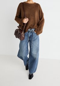 Pull en tricot brun, jean bleu clair à coupe ample avec cordon de serrage, associé à des bottines noires et un petit sac à main brun.