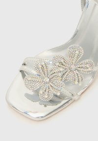 Ασημί πέδιλο με ψηλό τακούνι, με γυαλιστερό φινίρισμα, δύο ανθικά διακοσμητικά με πολύχρωμες στρας, και τετράγωνη μύτη.