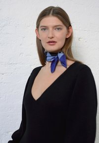 Suéter preto com decote em V e uma textura suave, combinado com um cachecol estampado em azul e roxo amarrado ao pescoço, sobre um fundo branco.