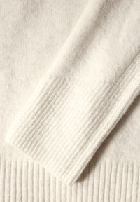Details eines hellen, gestrickten Pullovers mit gerippten Bündchen und einer weichen Stofftextur, die die einheitliche, glatte Oberfläche betont.