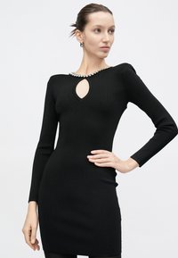 Γυναίκα πόζα σε εφαρμοστό μαύρο φόρεμα με μακριά μανίκια, κόψιμο σε σχήμα δακρύου στο ντεκολτέ και διακοσμητική κοσμημένη γιακά απέναντι σε απλό φόντο.