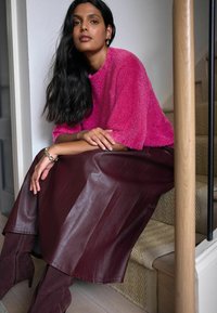 Helles pinkes, flauschiges Sweatshirt, burgunderrote Lederrock und passende, hochhackige Stiefel. Model sitzt an einer Treppe mit gewobener Textur im Hintergrund.