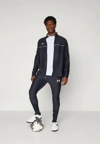 Juodas sportinis treningas su baltais akcentais, kuris apima džemperį užtrauktuku ir priglundančias kelnes. Modelis stovi ant futbolo kamuolio, dėvėdamas baltus sportinius batelius.