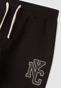 Czarne dresy z żeberkowym paskiem w talii, białymi sznurkami do ściągania oraz nadrukowanym logo "NY" w czerni i bieli na lewym udzie.