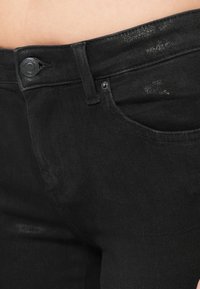 Fekete farmernadrág, amelyen a derékpánt, a gomb és a zseb közelképe látható. Az anyag enyhe fényű és texturált felületű.