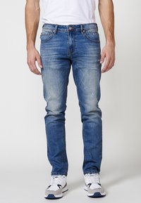 Jeans ajustados de color azul con un lavado desgastado, que cuentan con cinco bolsillos, presillas para cinturón y detalles de costura visibles. Combinados con zapatillas blancas.