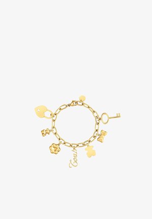 Złota bransoletka z łańcuszka z zawieszkami, w tym kłódką w kształcie serca, kluczem, misiem, kokardą, kwiatem i nazwą „Tous” pisaną kursywą.