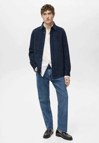 Navy blue shirt jacket with a collar, two chest pockets, and button closures, paired with light blue jeans and black loafers.