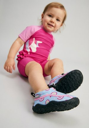 Kleinkind trägt pinke Shorts und ein Shirt, sitzt und trägt blaue Wasserschuhe mit geschlossener Spitze und pinken sowie regenbogenfarbenen Mustern vor hellem Hintergrund.